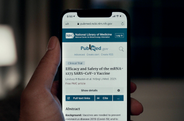 A hand holding a phone with the website PubMed reading "Efficacy and Safety of the mRNA-1273 SARS-CoV-2 Vaccine"