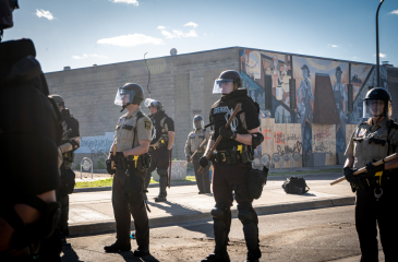 Police in a standoff with riot gear on and an urban landscape behind them