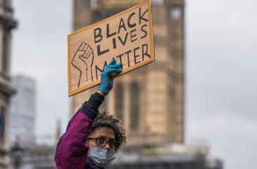 A Black woman with a mask on holds up a sign reading "Black Lives Matter" on it.