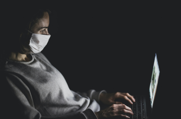 Person with a mask on staring at a laptop screen in the dark