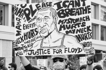 A black and white photo of a man in a mask holding a protest sign with George Floyd's face on it reading "Mama please, everything hurts. They are going to kill me. I can't breathe. My stomach hurts. My neck hurts. Water. Justice for Floyd."