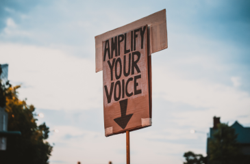 Cardboard protest sign held in the air reading "Amplify your voice" with an arrow down to the holder