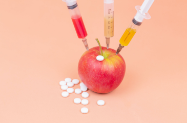 An apple with three syringes inserted into it and pills strewn about